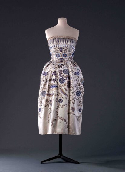 A strapless, knee-length cream-colored dress with intricate blue, silver, and gold floral embroidery, displayed on a mannequin. The bodice features a band of geometric patterns.
