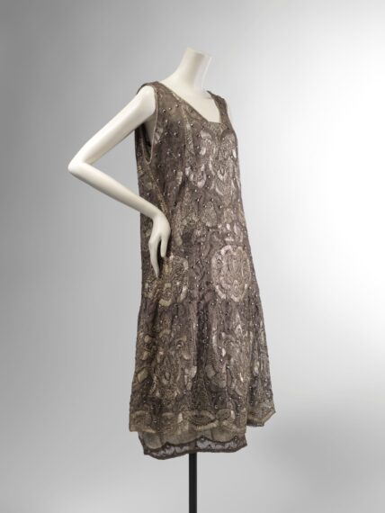 A sleeveless, short dress on a white mannequin, viewed from the back. The dress is heavily embroidered and beaded with intricate, swirling patterns in muted metallic tones. A wide, metallic ribbon is tied in a bow at the upper back, creating a V-shaped neckline.