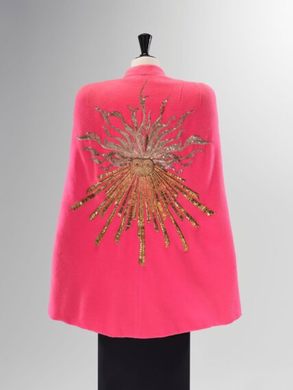 A close-up of the back of a bright pink wool cape. A large, stylized sun design is embroidered in gold sequins and thread, with a face at its center and rays extending outward.