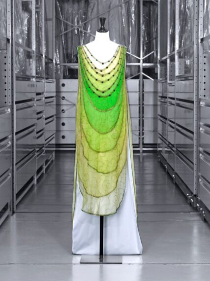 A layered, ombre green dress with a dropped waist displayed on a mannequin. The dress features beaded drapes and embroidered swirling patterns.