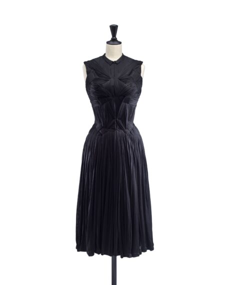 A black sleeveless, knee-length dress with a structured, pleated bodice and a flowing pleated skirt, displayed on a mannequin.