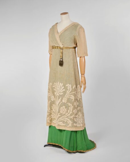 The image shows a mannequin dressed in a beige layered gown with half sleeves and a knee-length overlay featuring intricate white floral patterns on top of a green sheath.