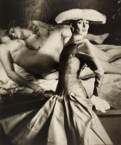 Black-and-white photo of a woman in a dress with a fitted bodice that flares out into a full skirt and hat with a blurred nude figure in the background.