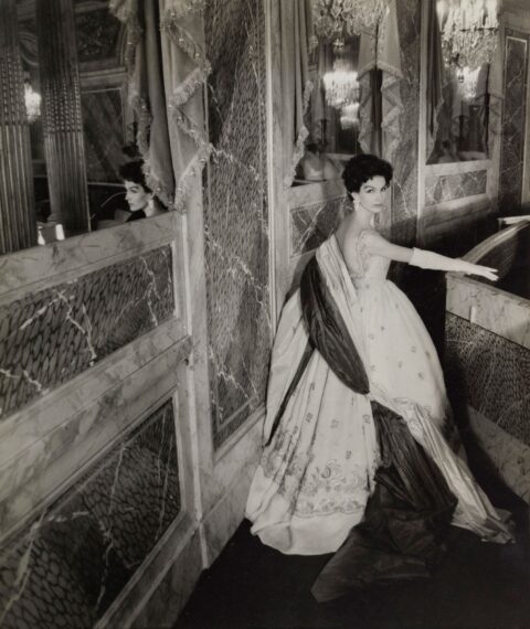 Black-and-white photo of a woman wearing an elegant gown standing in a lavishly decorated corridor.