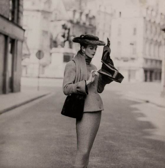 A black and white photograph shows a woman wearing a tailored coat with a high collar and a matching pencil skirt standing in the middle of a cobblestone street.