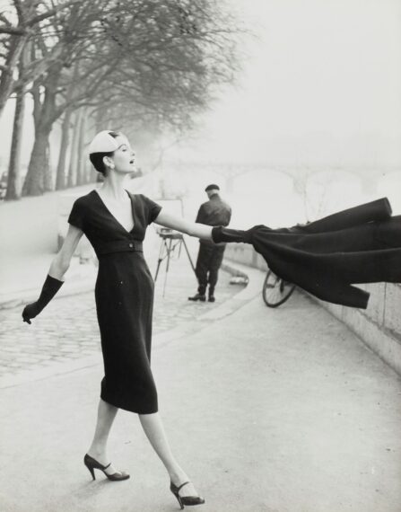 A black-and-white photograph shows a woman elegantly dressed in a fitted, dark-colored dress walking along a riverbank with trees.