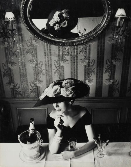 Black-and-white photo of a woman wearing an off-the-shoulder dress and a large hat adorned with flowers sitting at a table with a champagne bucket.