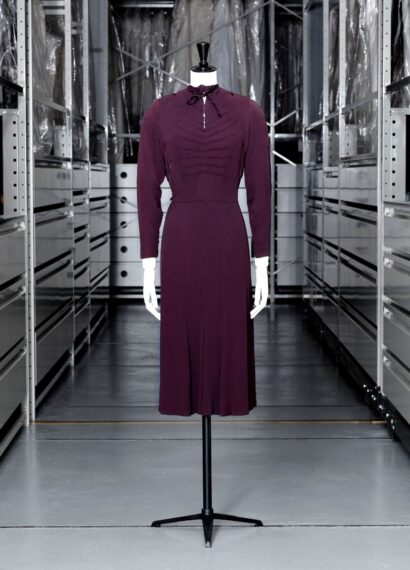 The image shows a mannequin wearing a dark purple dress, with long sleeves and a high neck, standing in a storage room.