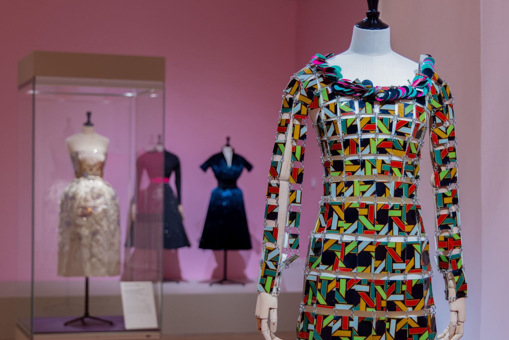 Colorful geometric long-sleeved mini dress on a mannequin in pink room with three other dresses displayed in the background.