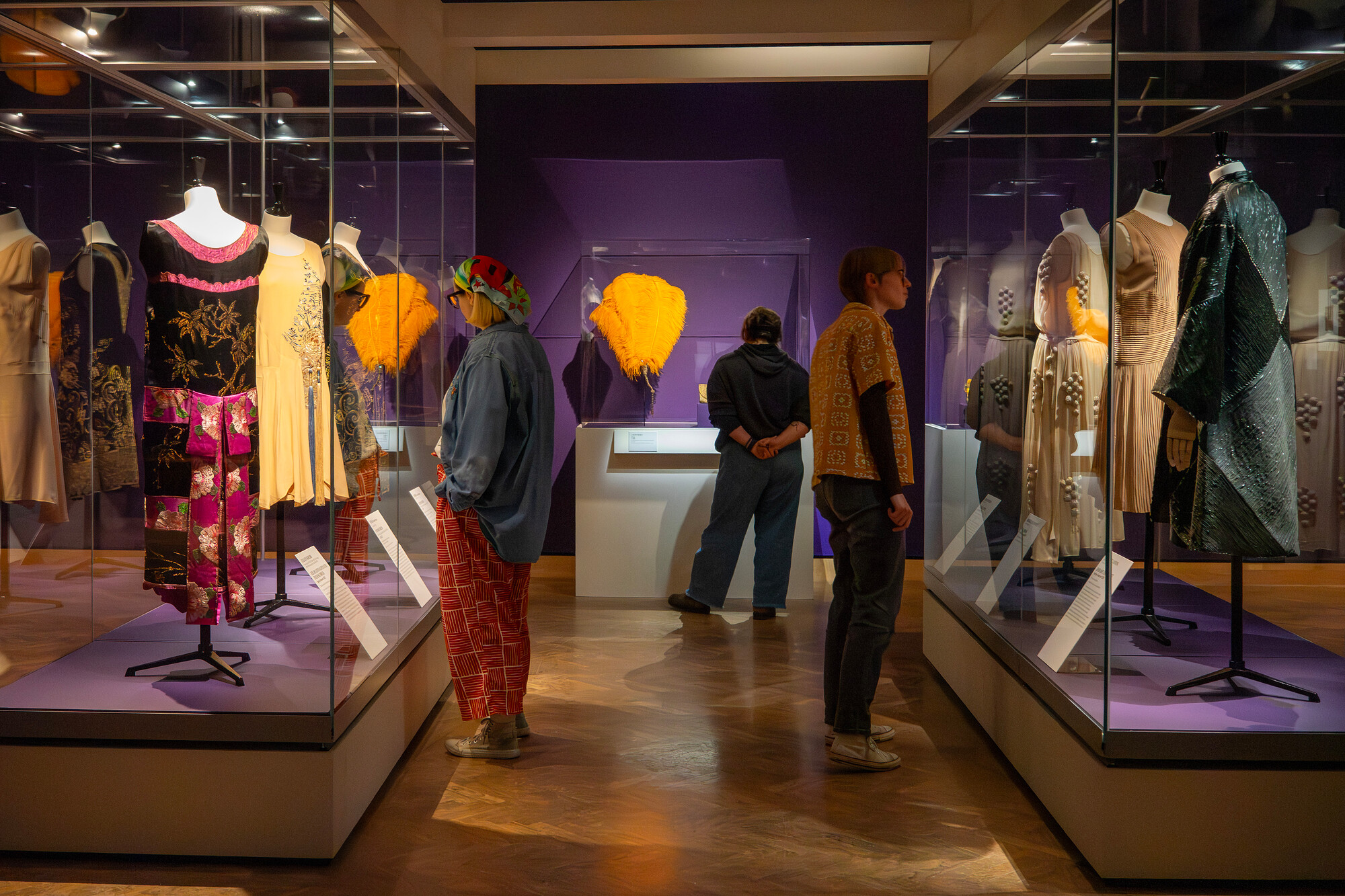The image shows museum visitors viewing an exhibition with dresses and other fashion pieces displayed in glass cases.