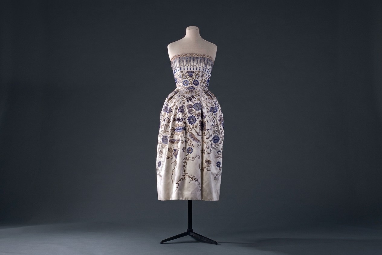 A strapless, knee-length cream-colored dress with intricate blue, silver, and gold floral embroidery, displayed on a mannequin. The bodice features a band of geometric patterns.